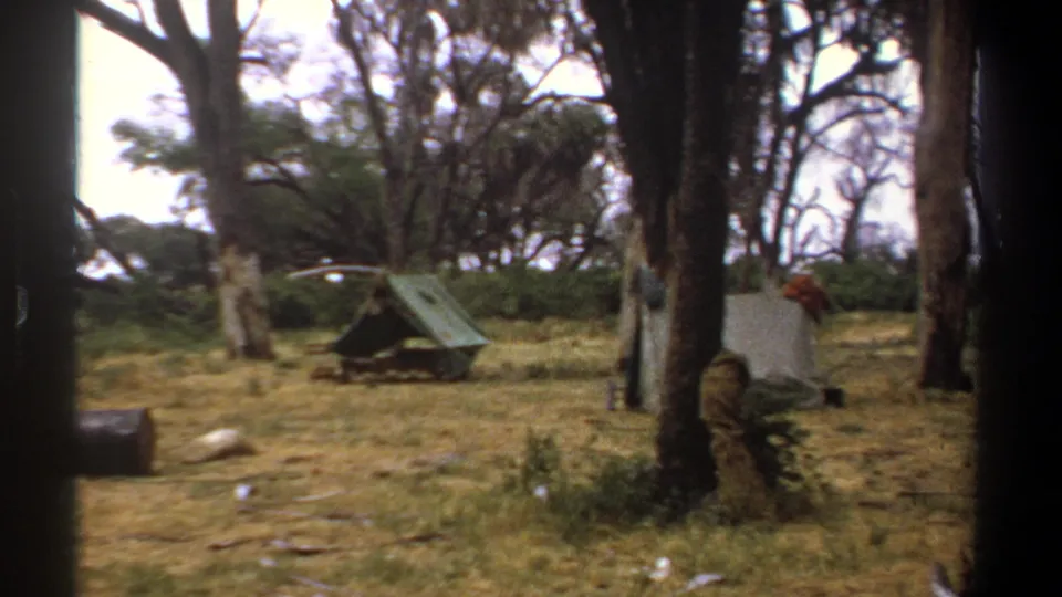 Kenya 1969 frame of a quiet wooded encampment with green and white tents set on dry grass behind dark viewfinder-like borders.