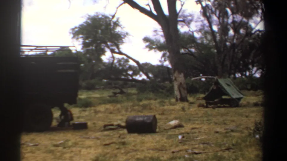 Kenya 1969 8mm frame with the side of a dark truck partly blocking a wooded camp where a green tent and ground containers are visible.