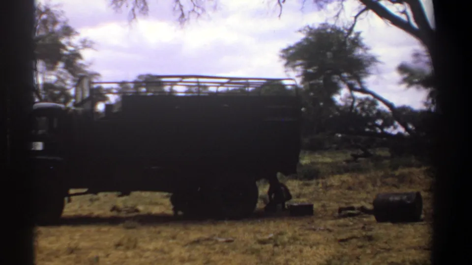 Kenya 1969 frame showing a dark utility truck with a roof rack parked in a wooded dry-grass encampment, with containers on the ground.