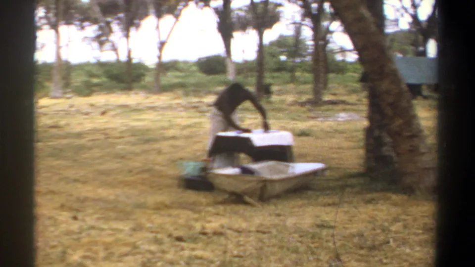 1969 Kenya 8mm frame showing a man working at a small white-covered table beside a wheelbarrow in a dry wooded clearing.