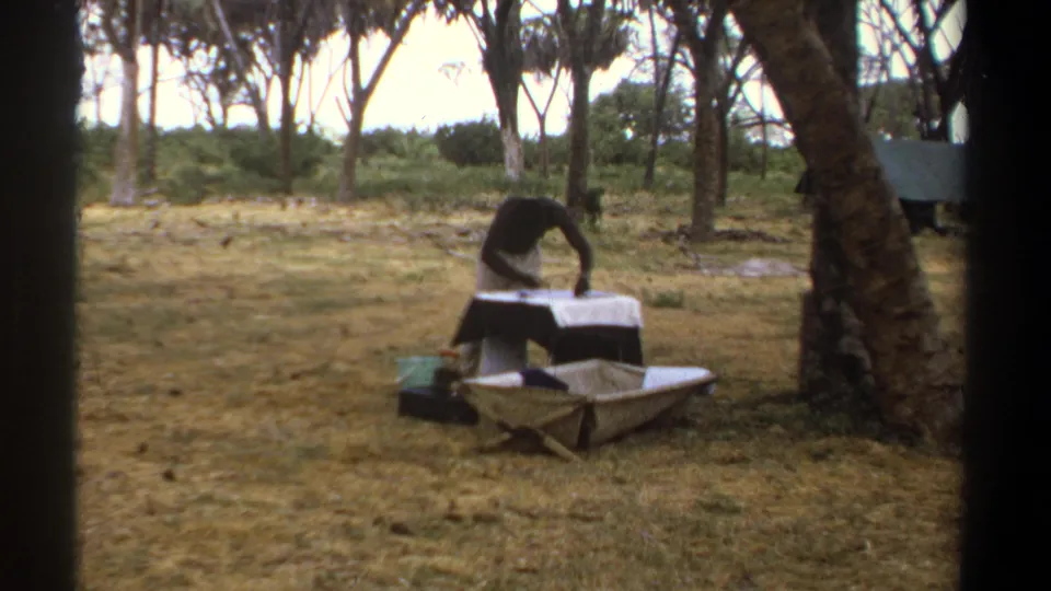 1969 Kenya home-movie frame of a man handling items on a white-covered table, with a wheelbarrow and trees in the clearing.