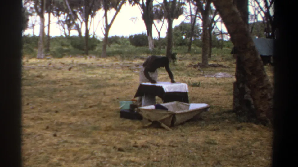 1969 Kenya 8mm frame showing the man still bent over the covered table, with the wheelbarrow in front and trees behind.
