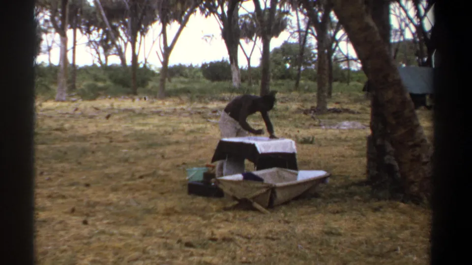 In Kenya 1969 home-movie footage, an adult man bends over a low table or work surface beneath trees in dry grass.