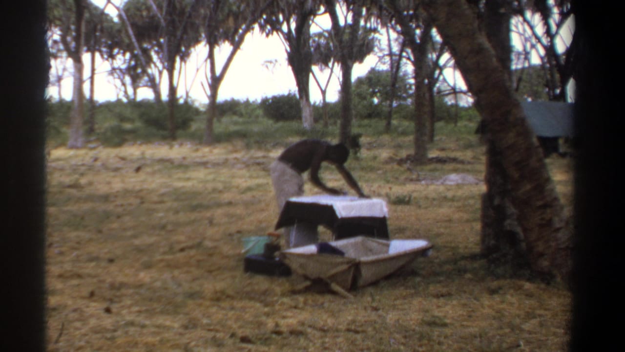 The man continues leaning over a pale metal-looking object on a table, with trees and dry grass visible around him in Kenya.