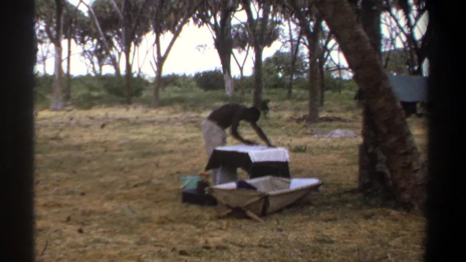 The man continues leaning over a pale metal-looking object on a table, with trees and dry grass visible around him in Kenya.
