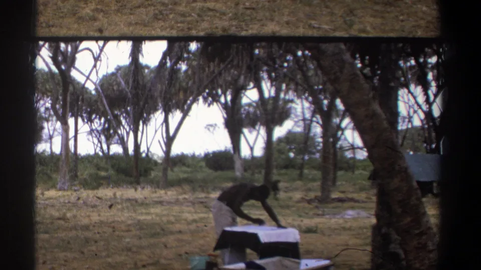 The final frame shows the man still bent over the table near a tree, continuing the outdoor cleaning or scraping task in Kenya.