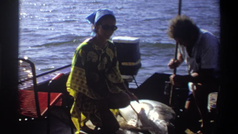 1969 Nigeria 8mm frame with a woman in a blue headscarf and patterned dress smiling near a large fish as another adult works beside it on the boat.