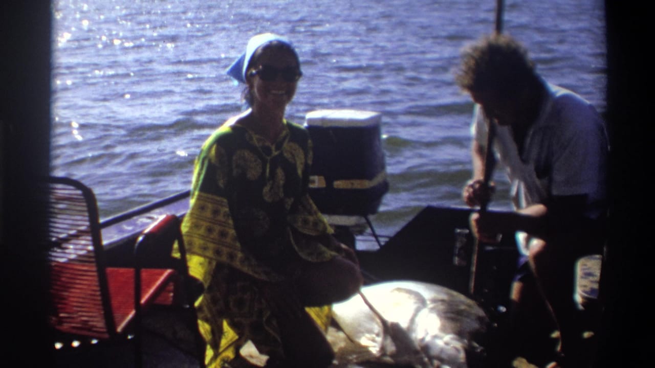 Woman in patterned clothing kneels beside a large fish on a small boat while a man works near the stern.