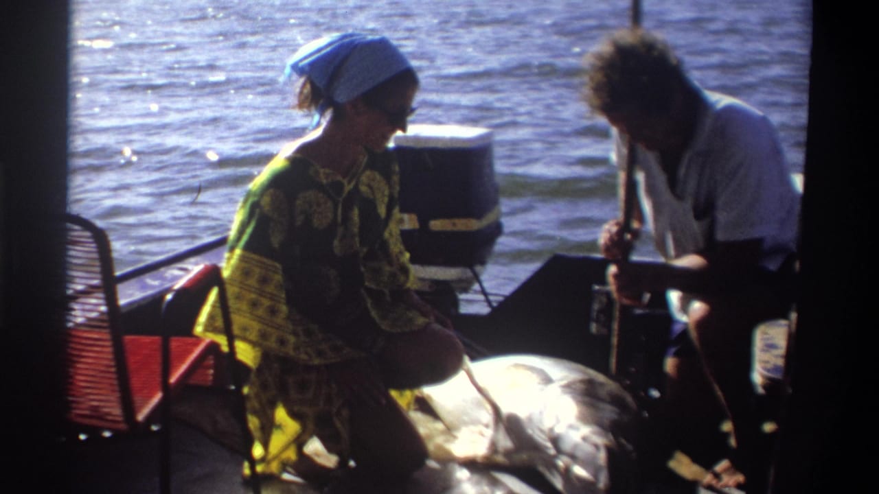 Woman in a blue headscarf kneels by a large fish as a man leans over gear at the stern of the boat.