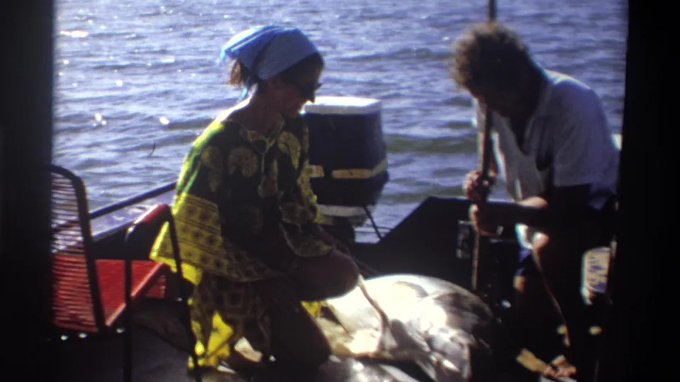 1969 Nigeria frame of two adults on a small motorboat, with the woman turned sideways near a large fish and bright water behind.