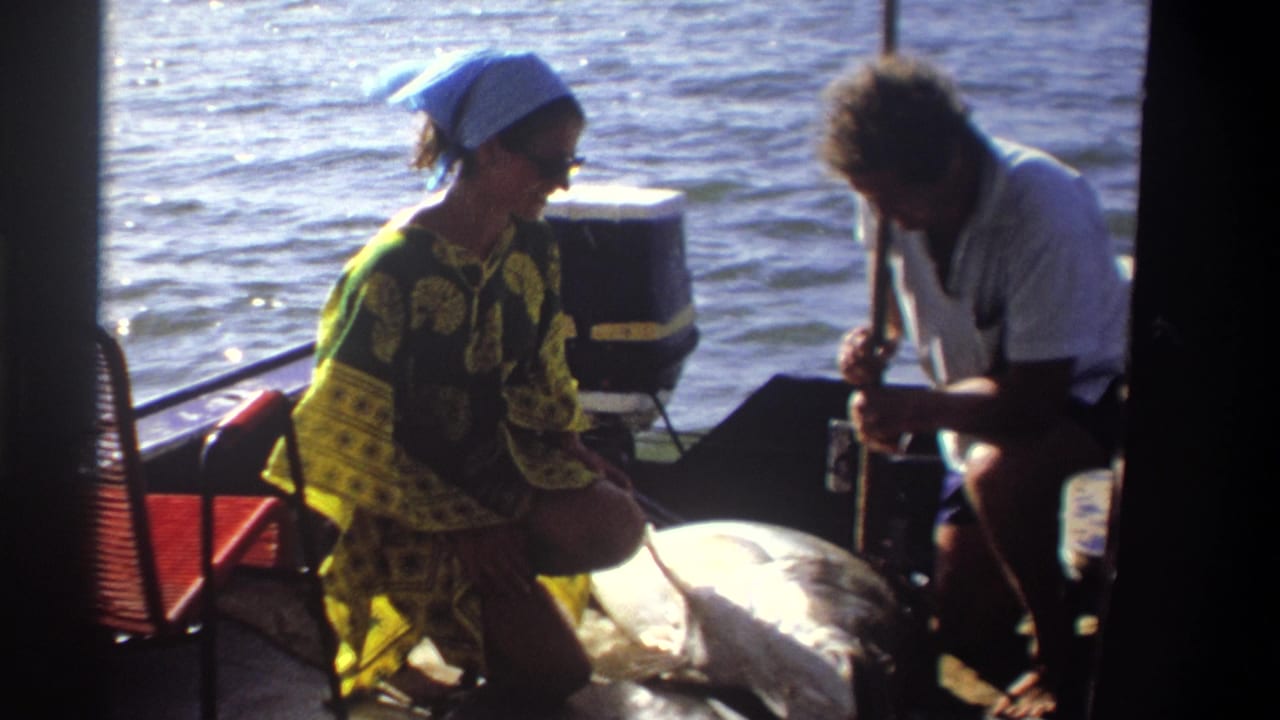 1969 Nigeria home-movie still with a woman kneeling beside a large fish while a man handles something near the stern.