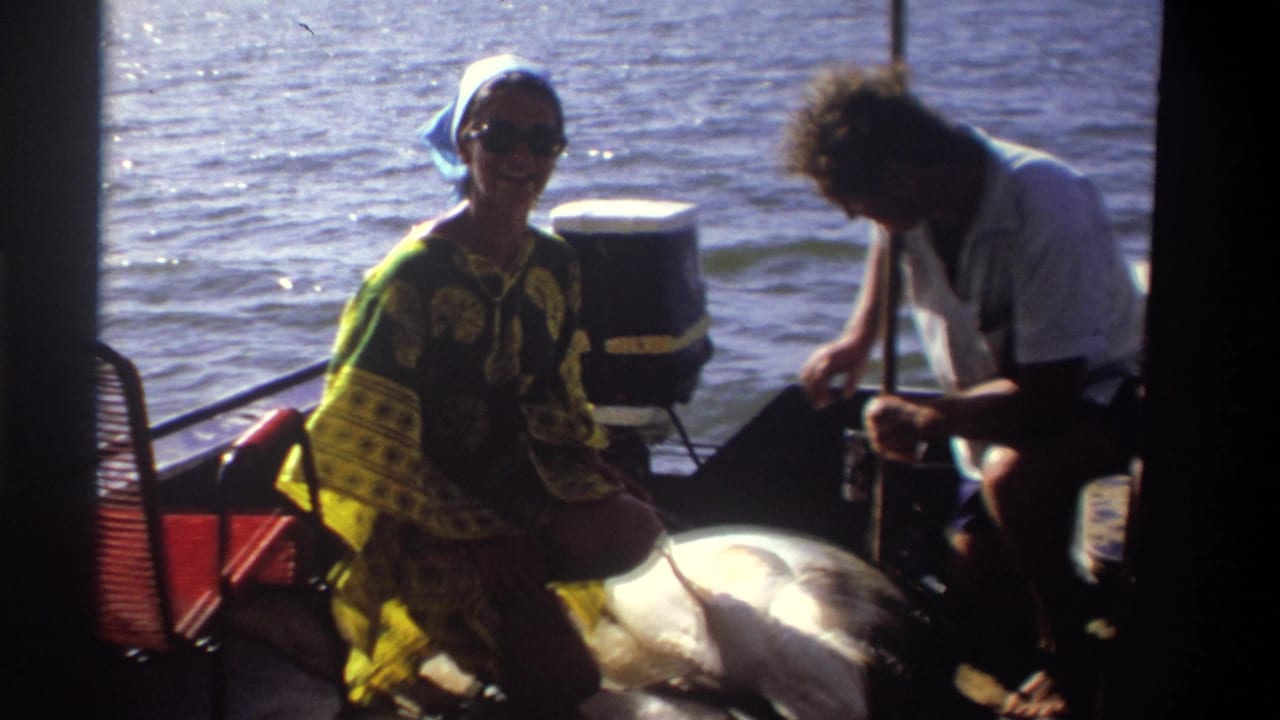 1969 Nigeria home-movie frame showing a woman smiling toward the camera beside a large fish on a small boat in bright sun.