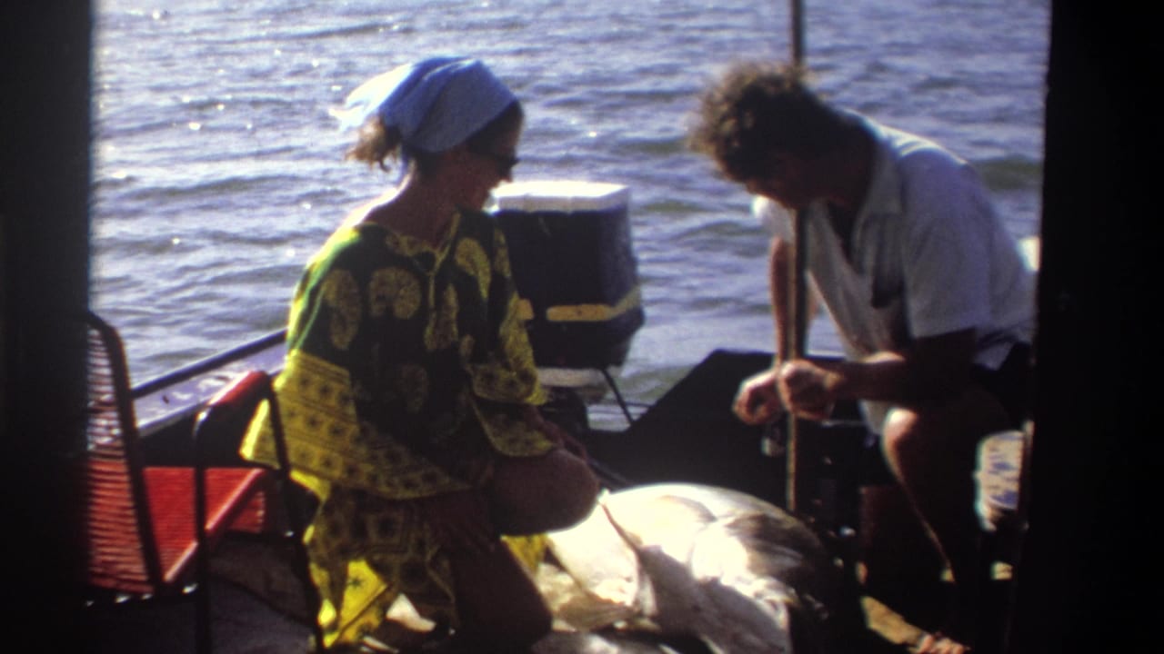 In 1969 Nigeria home-movie footage, two adults sit on a small boat as they examine a large fish near a cooler and red chair.