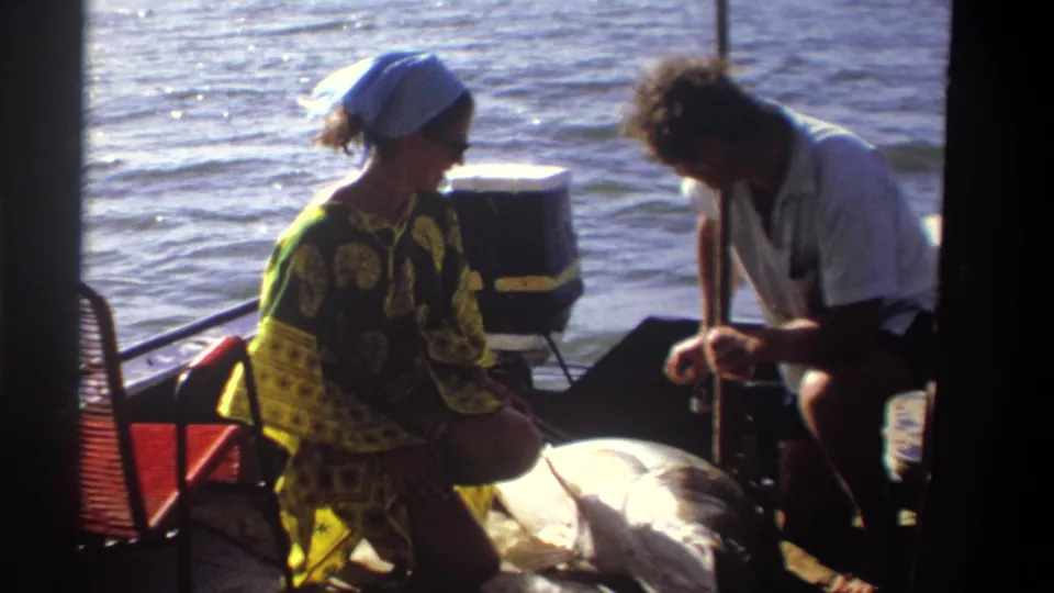 In 1969 Nigeria home-movie footage, two adults sit on a small boat as they examine a large fish near a cooler and red chair.