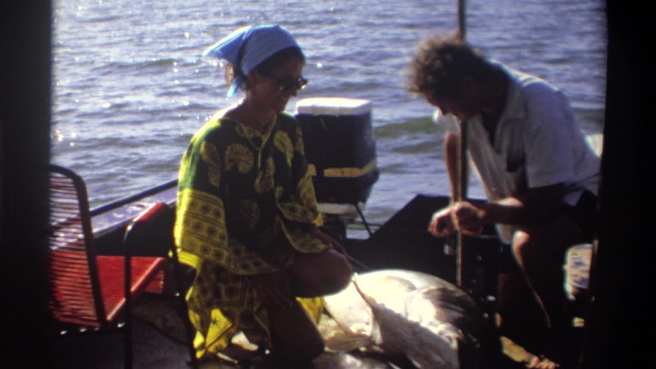 Woman in a blue headscarf sits near a large fish while a man crouches beside it.