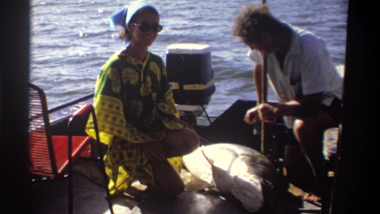 The woman smiles toward the 8mm camera during a 1969 Nigeria boating scene while the man remains focused on the large fish.
