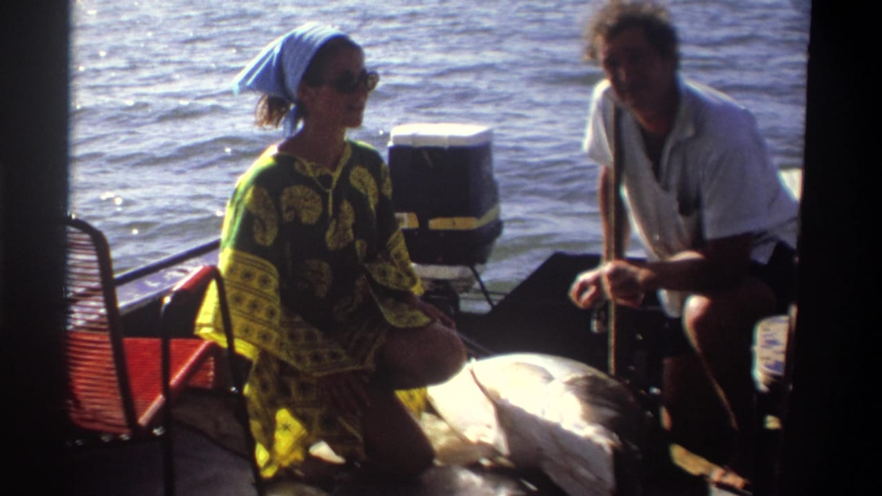 Two adults on a small boat in 1969 Nigeria face the camera, with a large fish lying in the foreground under bright sunlight.