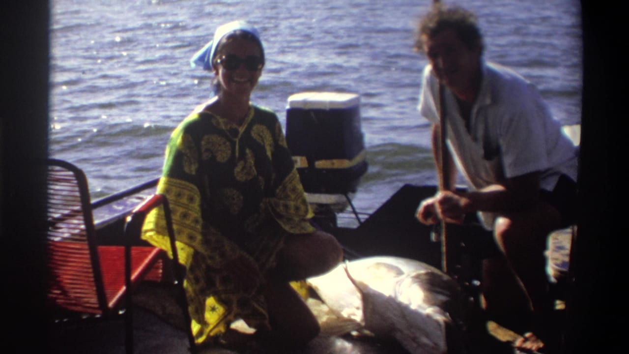 In Nigeria 1969 home-movie footage, two adults sit on a boat near open water, one wearing sunglasses and a patterned coverup.