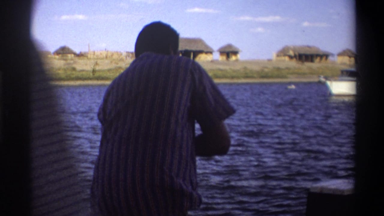 A man in a striped shirt stands on a boat in Nigeria 1969 footage, looking across water toward thatched structures on the shore.