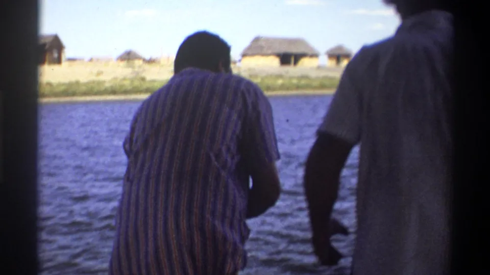 Two men stand on a boat in Nigeria 1969 footage, looking over the water toward a shoreline with several thatched huts.