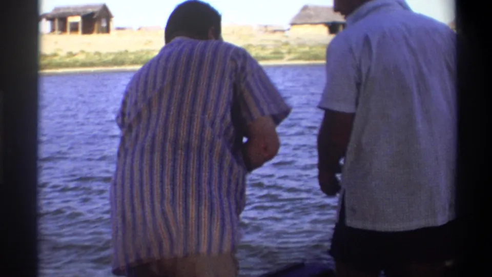 In Nigeria 1969 8mm footage, two men stand on a boat and face a lakeshore or riverbank with thatched structures beyond the water.