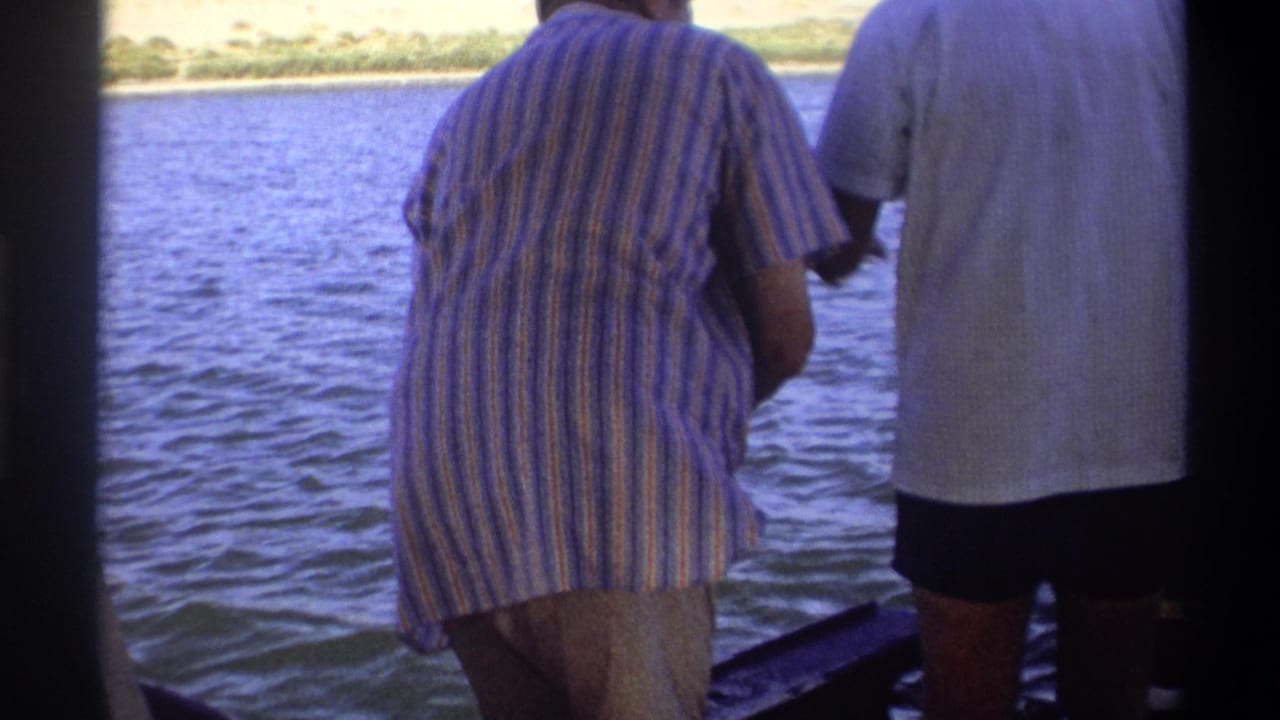 Two adult men in casual shirts stand at a boat rail on a Nigerian waterway, consistent with supplied 1969 home-movie metadata.