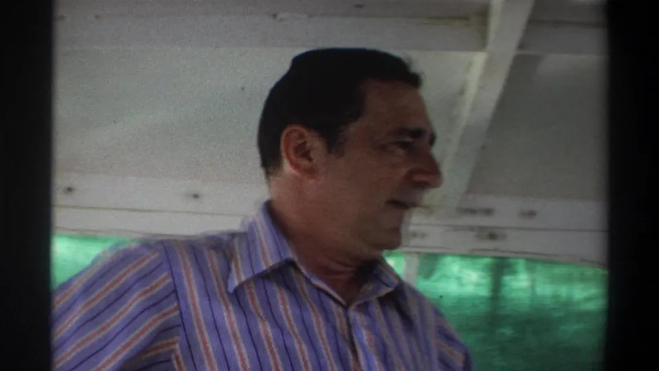 1969 Nigeria home-movie frame with a profile view of an adult man in a striped shirt under a canopy on a small boat.