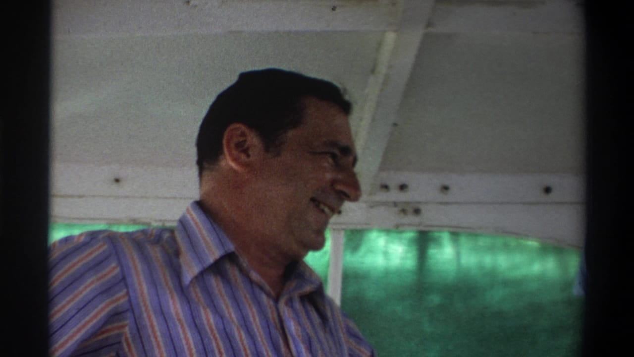 1969 Nigeria home-movie frame showing a man in a striped shirt inside a boat, with green water seen through the side opening.
