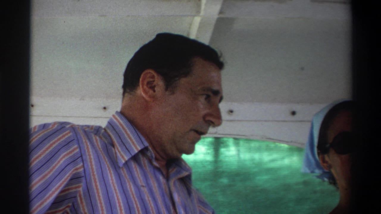 A man in a striped shirt looks down in the boat, with another passenger partly visible beside him in the 1969 Nigeria footage.