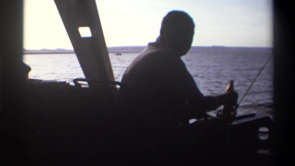 1969 Nigeria 8mm frame of a man in silhouette seated on a boat, with fishing pole, window frame, water, and horizon behind him.