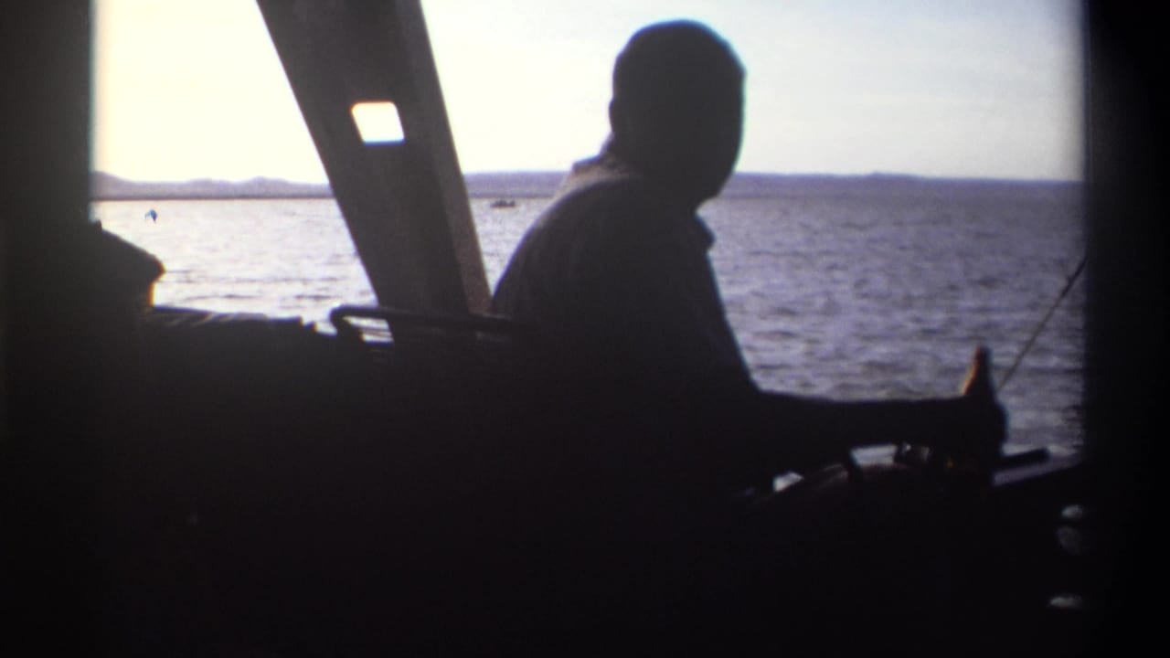 In Nigeria home-movie footage from 1969, a silhouetted man sits near the helm of a boat as water and low shoreline fill the window.