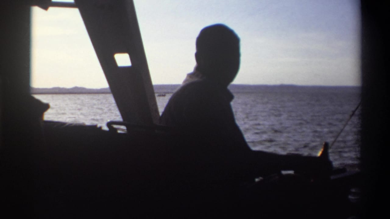 A silhouetted figure is seen from inside a boat in 1969 Nigeria, framed by angled supports with calm water and shoreline outside.