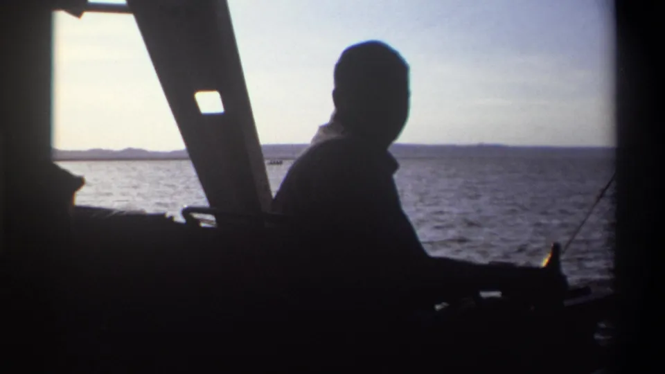A silhouetted figure is seen from inside a boat in 1969 Nigeria, framed by angled supports with calm water and shoreline outside.