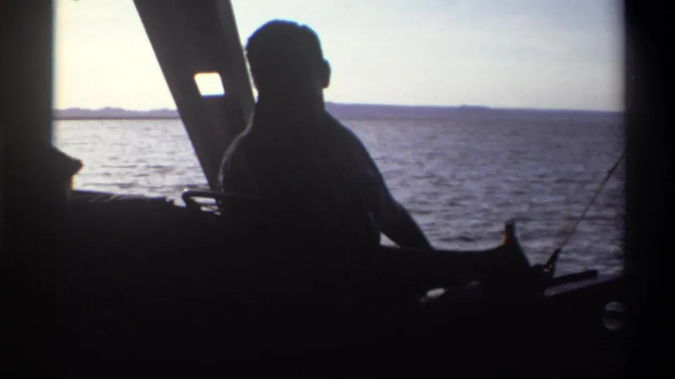 A man in silhouette turns inside the boat, with Nigeria's open water and distant shoreline visible through the bright window in 1969 footage.