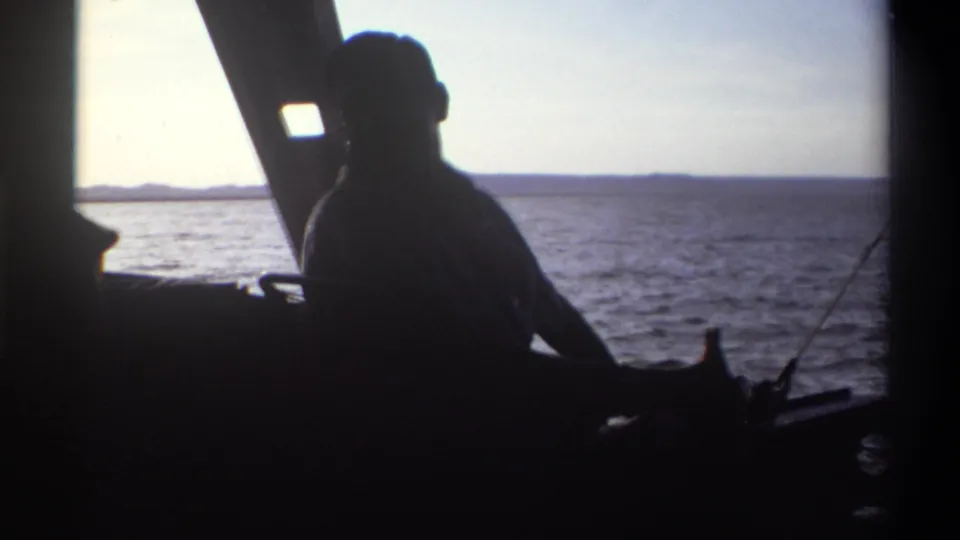 1969 Nigeria home-movie frame showing a silhouetted man seated aboard a boat against bright sea and distant land.