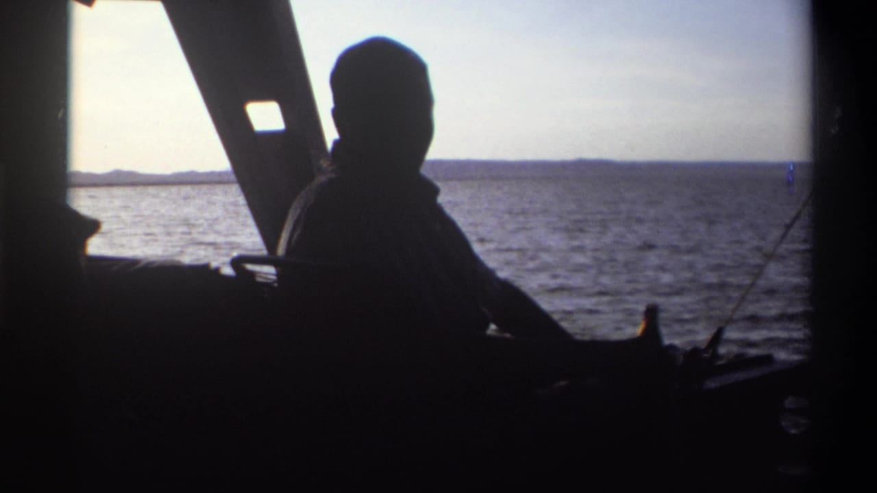 Silhouetted man aboard a boat in 1969 Nigeria, seated near a bottle with open water and shoreline in the background.