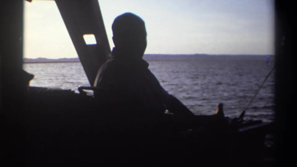Silhouetted man aboard a boat in 1969 Nigeria, seated near a bottle with open water and shoreline in the background.