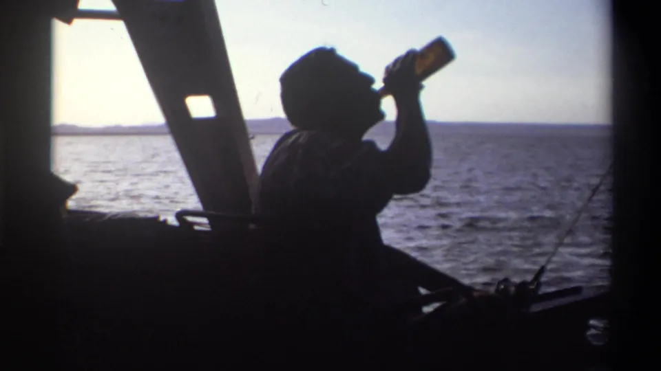 A 1969 Nigeria home-movie frame shows a silhouetted man drinking from a bottle aboard a boat against the sea horizon.