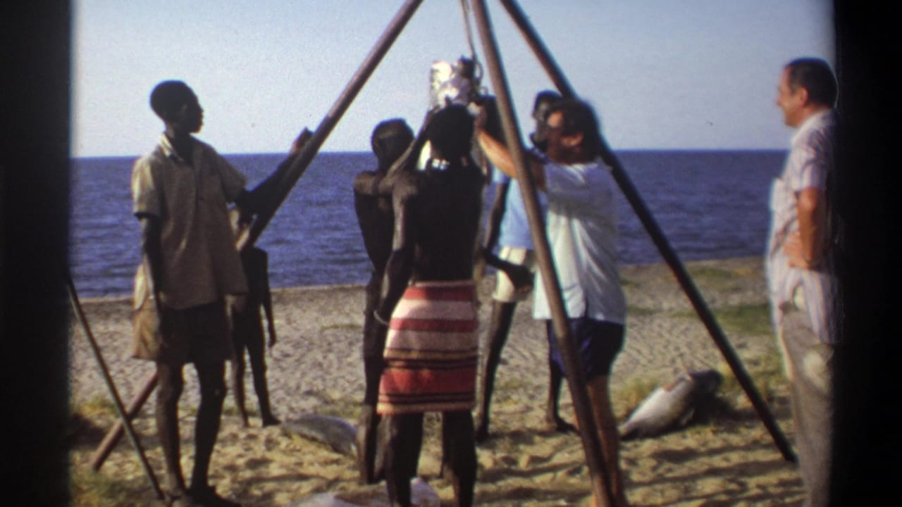 1969 Nigeria 8mm frame showing people gathered tightly around a suspended fish, with tripod poles and ocean visible behind them.