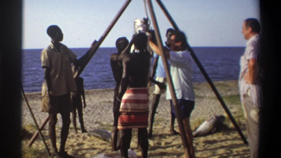1969 Nigeria 8mm frame showing people gathered tightly around a suspended fish, with tripod poles and ocean visible behind them.