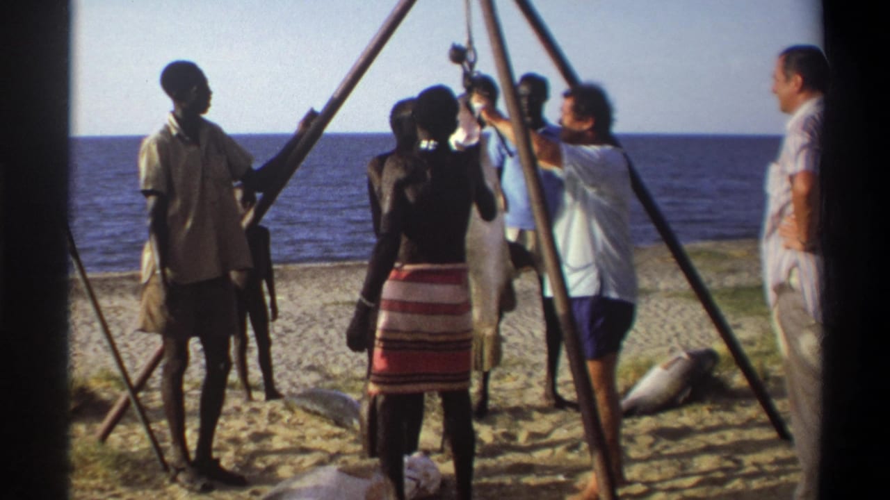 1969 Nigeria home-movie frame of adults beside a wooden beach hoist, with a large fish raised near the ocean horizon.