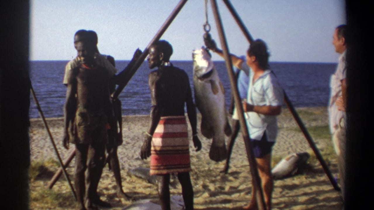 1969 Nigeria 8mm frame showing local men and Western visitors near a large suspended fish on a sandy beach.