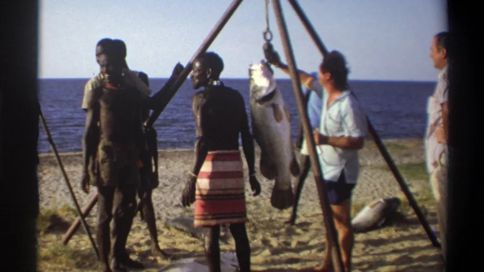 1969 Nigeria 8mm frame showing local men and Western visitors near a large suspended fish on a sandy beach.