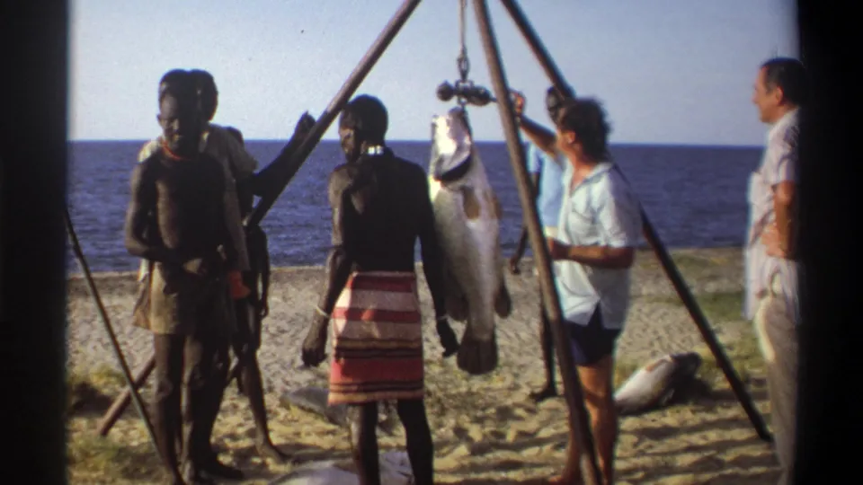 1969 Nigeria beach scene with a suspended fish, local men nearby, and visitors observing beside the shoreline.