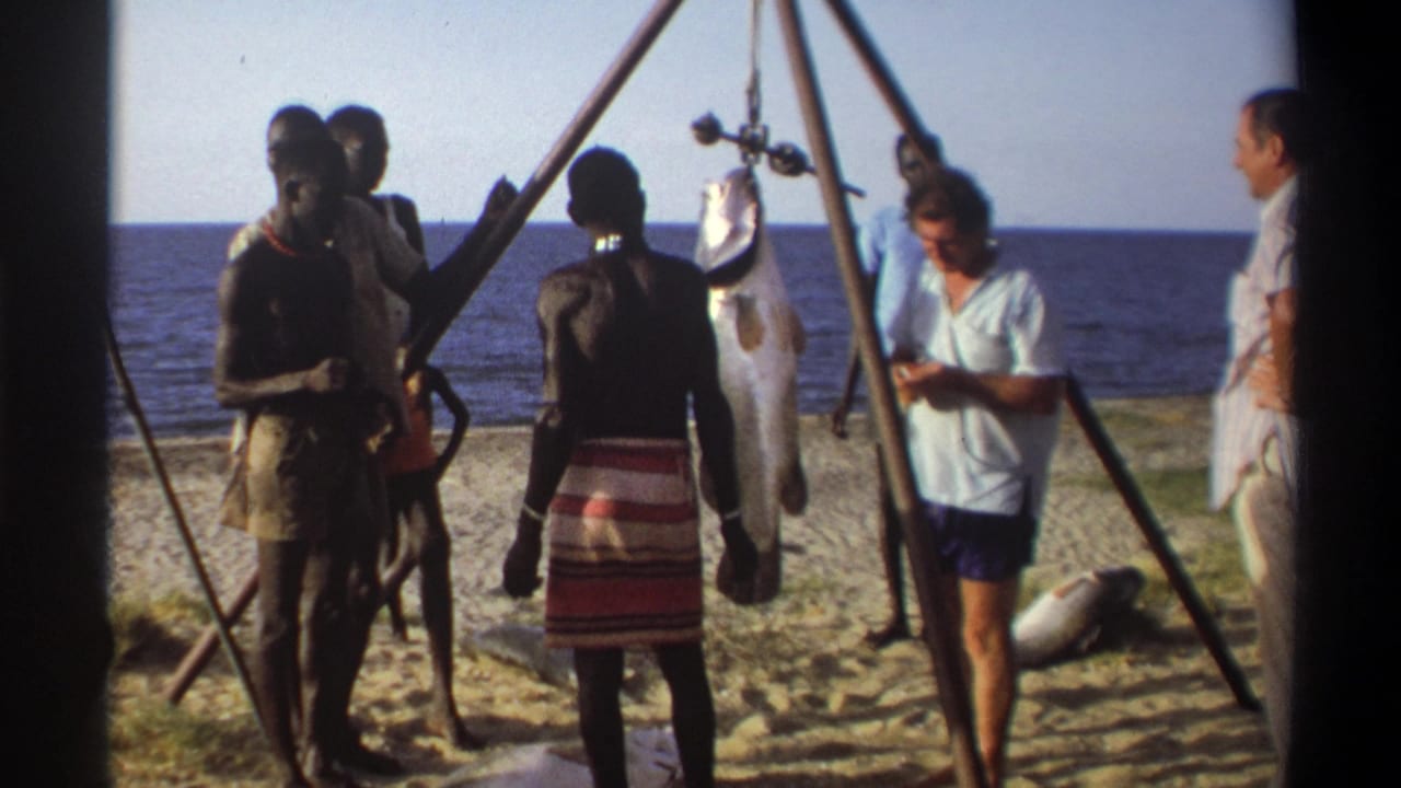 1969 Nigeria home-movie frame showing men and visitors around a tripod-held fish with the sea behind them.