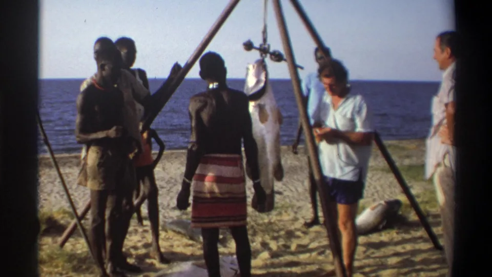 1969 Nigeria home-movie frame showing men and visitors around a tripod-held fish with the sea behind them.