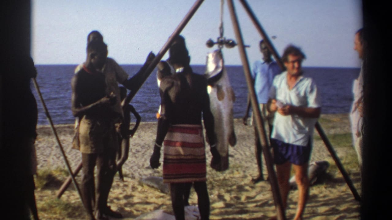 1969 Nigeria 8mm frame with local men, visitors, a suspended fish, and beach shoreline in bright daylight.
