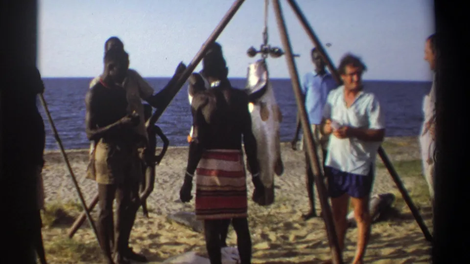 1969 Nigeria 8mm frame with local men, visitors, a suspended fish, and beach shoreline in bright daylight.