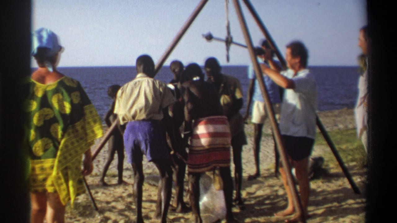 1969 Nigeria 8mm frame showing adults on a sandy shoreline around an A-frame scaffold with a large fish suspended by a hook.