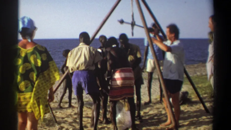 1969 Nigeria 8mm frame showing adults on a sandy shoreline around an A-frame scaffold with a large fish suspended by a hook.